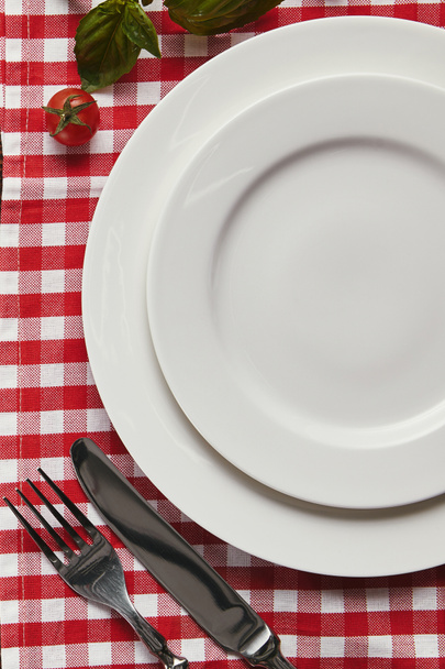 vista superior de pratos brancos vazios, talheres e manjericão fresco com tomate na toalha de mesa quadriculada
 - Foto, Imagem