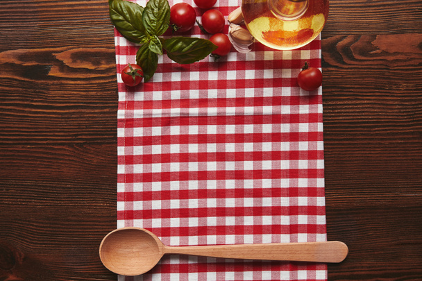 pohled shora kostkovaný ubrus, dřevěnou lžící a čerstvou bazalkou, rajčaty a olejem na dřevěné ploše - Fotografie, Obrázek