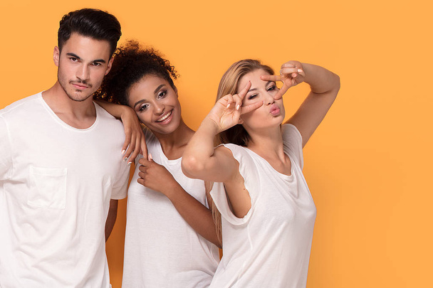 Three young multi ethnic people smiling to the camera, posing on orange studio background in white casual shirts. - Photo, Image
