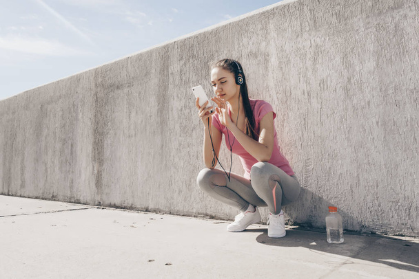 cute girl in pink t-shirt resting after training and jogging outdoors, listening to music on headphones - Фото, зображення