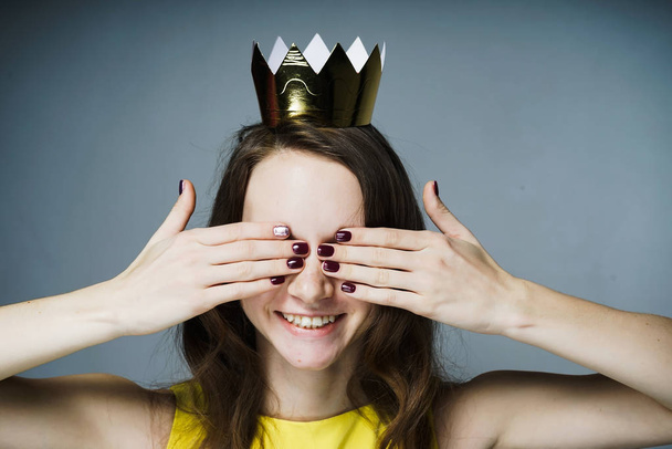 a cute smiling girl in a yellow dress covered her eyes with her hands, a golden crown on her head - Foto, afbeelding