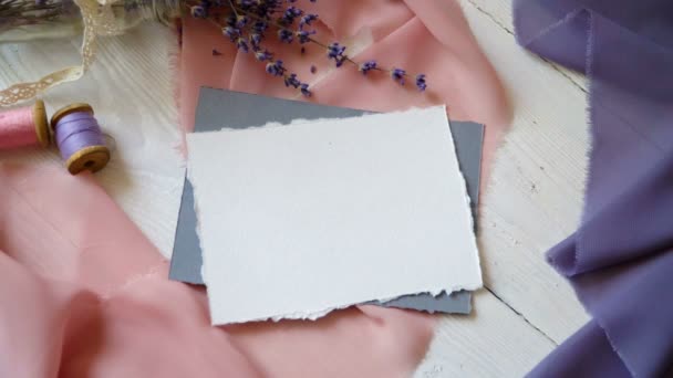 Scheda bianca, busta e nastro bianco su sfondo di tessuto rosa e blu con fiori di lavanda su sfondo bianco. Mockup con busta e carta bianca. Piatto. Vista dall'alto
. - Filmati, video