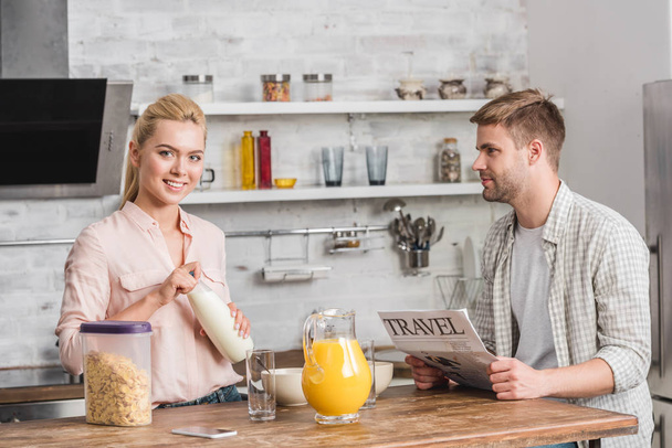 boyfriend holding newspaper and girlfriend opening bottle of milk during breakfast in kitchen - Фото, изображение