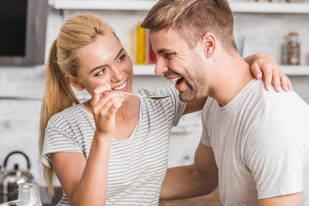 portrait of smiling girlfriend feeding handsome boyfriend with cornflakes in kitchen - Photo, Image