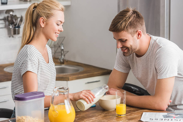 girlfriend pouring milk from bottle into boyfriend plate with cornflakes in kitchen - Foto, Imagen