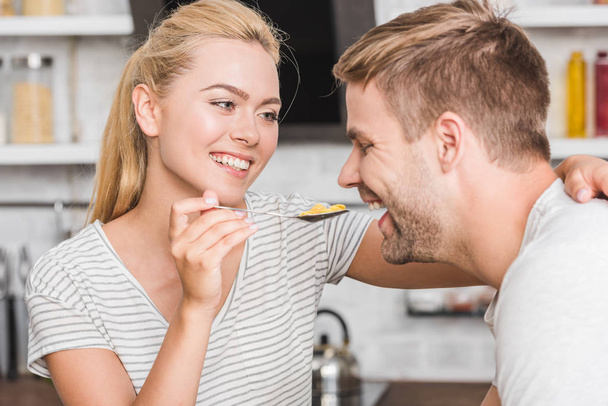 portrait of smiling girlfriend feeding boyfriend with cornflakes in kitchen  - 写真・画像