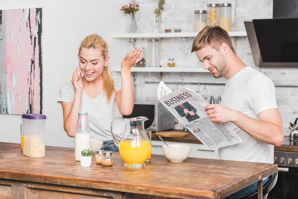 girlfriend talking by smartphone and gesturing during breakfast in kitchen, boyfriend reading newspaper - 写真・画像