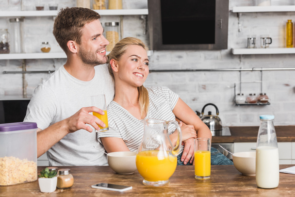 feliz novio abrazando novia durante el desayuno y sosteniendo el vaso de jugo en la cocina
 - Foto, Imagen