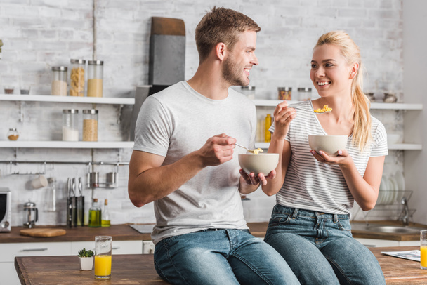alegre pareja sosteniendo platos y cucharas con copos de maíz y mirándose en la cocina
 - Foto, imagen