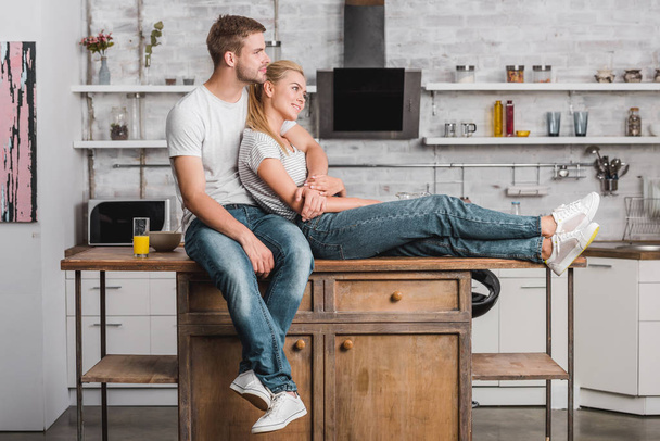 happy couple hugging, looking away and sitting on kitchen counter - Фото, зображення
