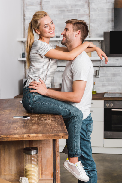 happy girlfriend sitting on kitchen counter and cuddling boyfriend - Photo, Image