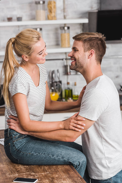 happy girlfriend sitting on kitchen counter and looking at boyfriend - Фото, изображение