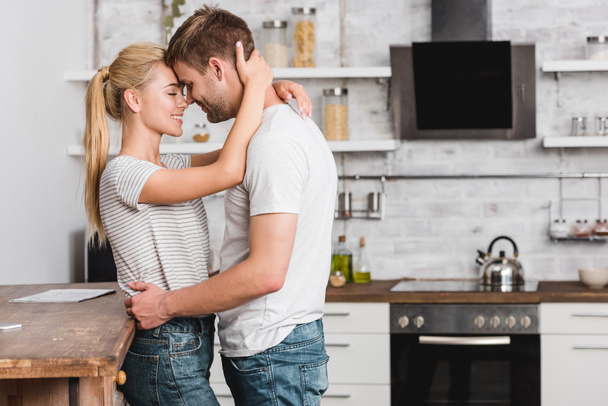 side view of couple cuddling in kitchen and leaning on kitchen counter - Foto, Imagem