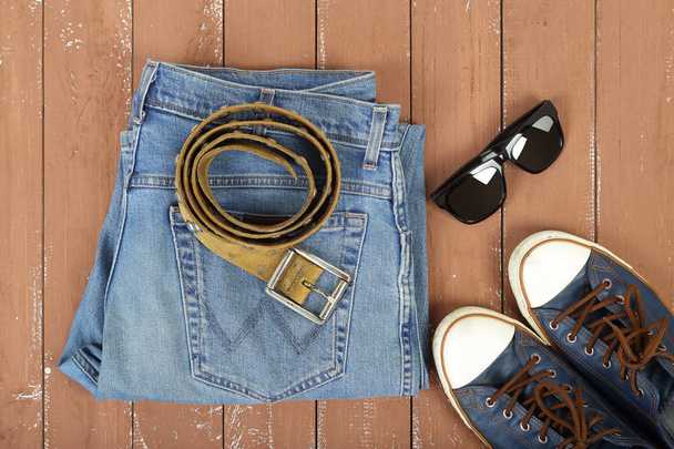 Clothes, shoes and accessories - Top view sunglasses, leather belt, gumshoes shoes and blue jeans on a wooden background - Zdjęcie, obraz