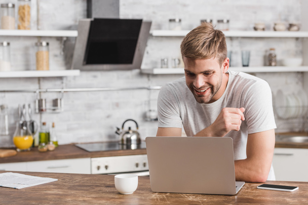 smiling handsome freelancer looking at laptop in kitchen - Фото, изображение