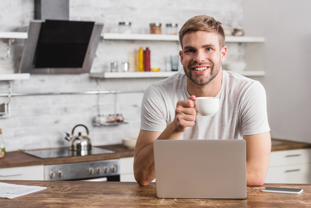 handsome smiling man holding cup of coffee and looking at camera in kitchen - Foto, Bild