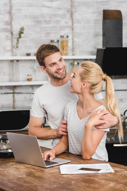 novio sonriente abrazando novia en la cocina mientras ella trabaja con el ordenador portátil
 - Foto, Imagen