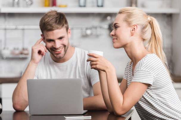 smiling couple sitting in kitchen with laptop and cup of coffee - Foto, Bild