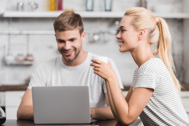 smiling young man using laptop while happy girlfriend drinking coffee - Photo, Image