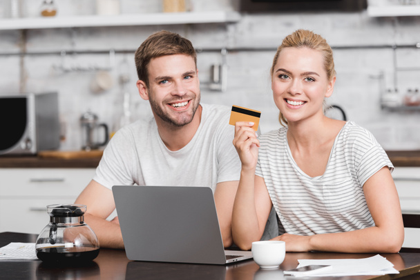 heureux jeune couple avec carte de crédit en utilisant un ordinateur portable et souriant à la caméra à la maison
 - Photo, image