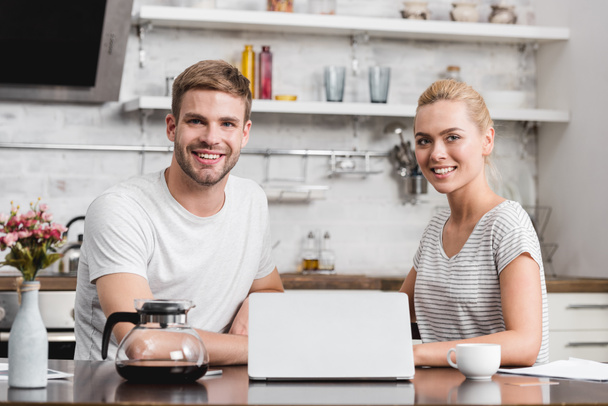 happy young couple using laptop and smiling at camera in kitchen - Foto, Imagem