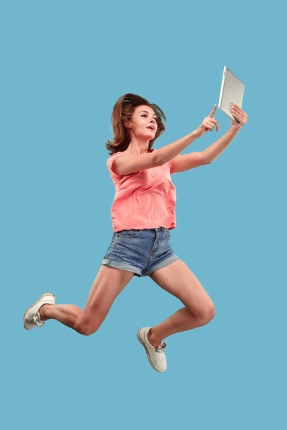 Image of young woman over blue background using laptop computer or tablet gadget while jumping. - Φωτογραφία, εικόνα