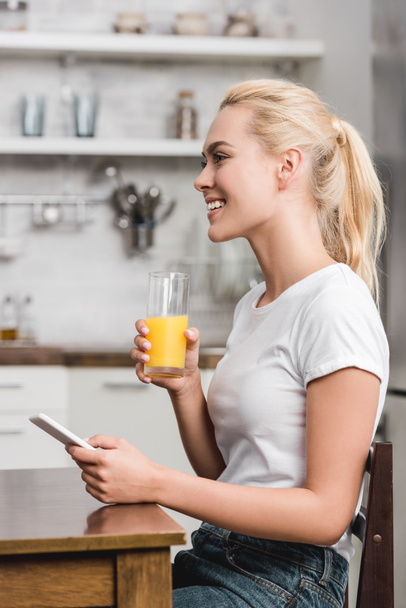 side view of smiling young woman using smartphone and holding glass with fresh juice - Foto, imagen