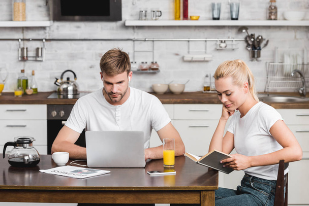 молодой человек с ноутбуком и улыбающаяся женщина читают книгу, сидя вместе за кухонным столом
 - Фото, изображение