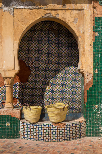 Non functional water tap on the street in Rabat, Morocco. Geometric pattern of the colorful traditional mosaic. Two baskets on the small wall. Weathered wall with an arch and column. - Photo, Image