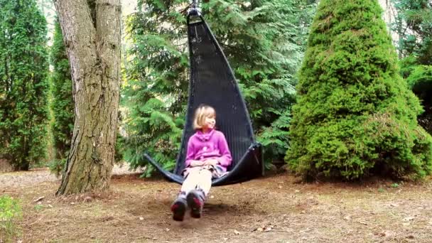 Little beautiful girl on black rocking chair hangs on tree branch in wooded homestead. - Footage, Video
