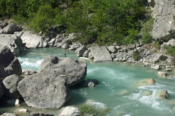 Klarer blassblauer, schnell fließender Fluss mit großen Felsen und Stromschnellen in der Nähe des Dorfes rimon im Dromtal, Frankreich. Bäume säumen den Fluss als Hintergrund. - Foto, Bild