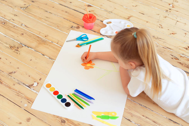 Little blonde girl painting on big white paper while laying on floor indoors - Foto, afbeelding