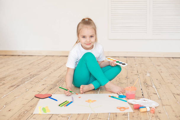 adorable girl painting on big white paper and looking at camera while sitting on wooden floor at home - Фото, зображення
