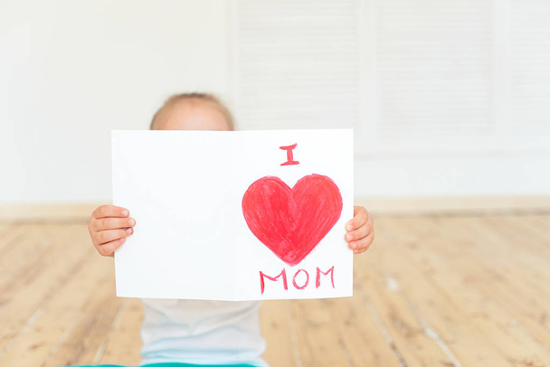 Little girl holding Mom greeting card in front of face while sitting on floor , Mothers Day concept - Photo, Image