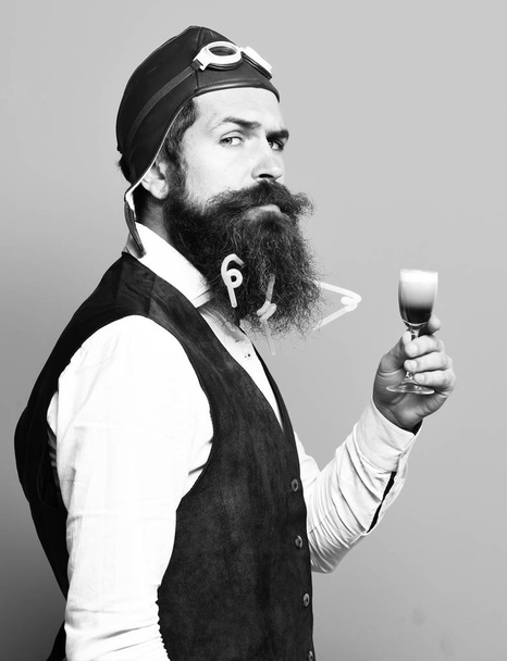 handsome bearded pilot or aviator man with long beard and mustache on funny face holding glass of alcoholic shot in vintage suede leather waistcoat with hat and glasses on blue studio background - Fotó, kép