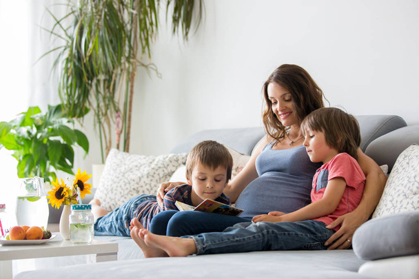Młoda kobieta w ciąży, czytanie książki w domu do jej dwóch chłopców, jedzenie owoców, przytulanie i śmiejąc się - Zdjęcie, obraz