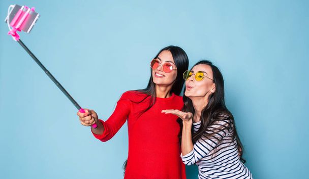 Portrait of two stylish brunettes in sunglasses making selfie using selfie stick and smartphone on blue background - Foto, afbeelding