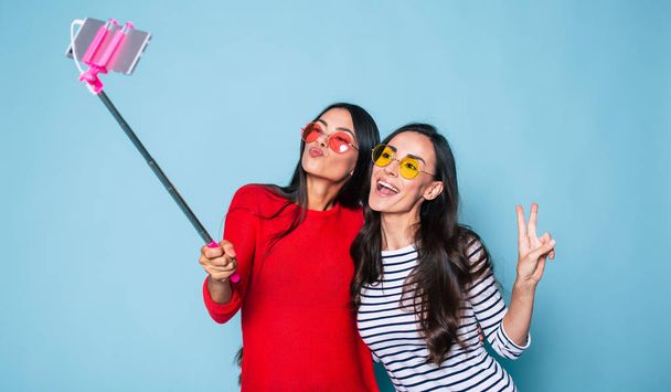 Portrait of two stylish brunettes in sunglasses making selfie using selfie stick and smartphone on blue background - Foto, afbeelding