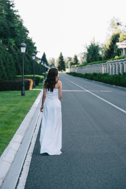 вид сзади женщины в белом свадебном платье, идущей по дороге
 - Фото, изображение