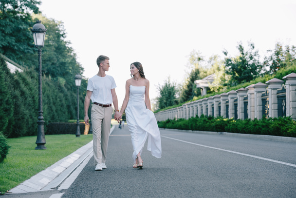 novia en vestido de novia y novio tomados de la mano y caminando por la carretera
 - Foto, Imagen