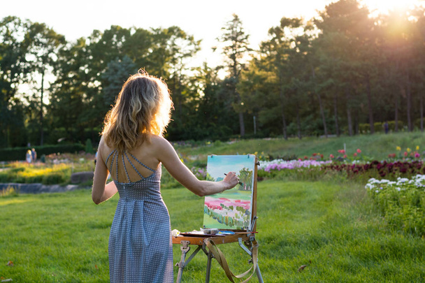 Una joven artista sostiene un pincel y pinta un cuadro en un caballete en los rayos de la puesta del sol. El pintor pinta pinturas al óleo en el jardín al atardecer
 - Foto, Imagen
