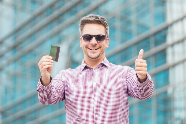 Portrait of young businessman holding credit card and showing thumbs up with office building in the background. - Fotoğraf, Görsel