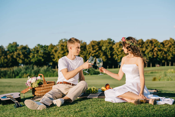 happy wedding couple clinking with glasses of wine during picnic on green lawn - Foto, immagini