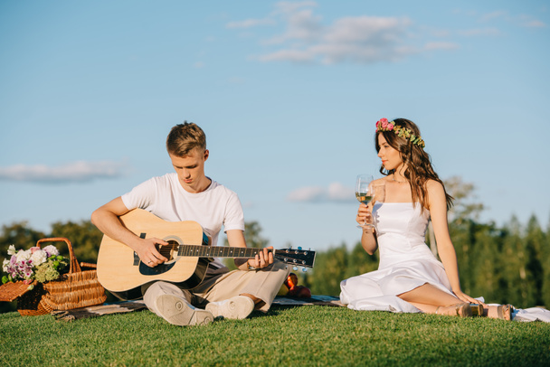 jóképű vőlegény játszik akusztikus gitár menyasszony borral romantikus piknik közben - Fotó, kép
