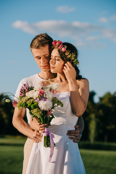groom embracing his attractive bride in flower wreath with wedding bouquet - Photo, Image