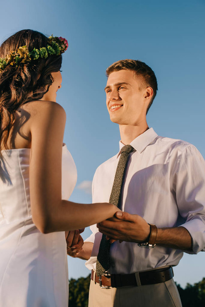Blick auf ein glückliches junges Hochzeitspaar, das sich an den Händen hält und vor blauem Himmel lächelt - Foto, Bild