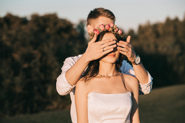 mladý ženich zavřel oči krásné usměvavé nevěstě v parku  - Fotografie, Obrázek