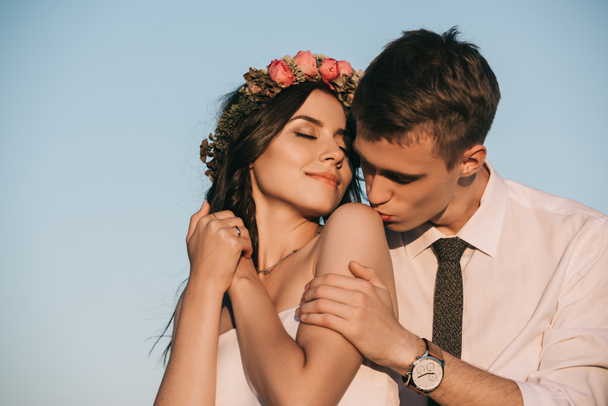 jovem noivo beijando bela noiva sorridente contra o céu azul
  - Foto, Imagem