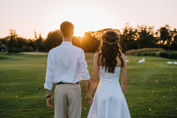 Rückansicht von jungen Hochzeitspaaren, die sich Händchen haltend im Park bei Sonnenuntergang stehen  - Foto, Bild