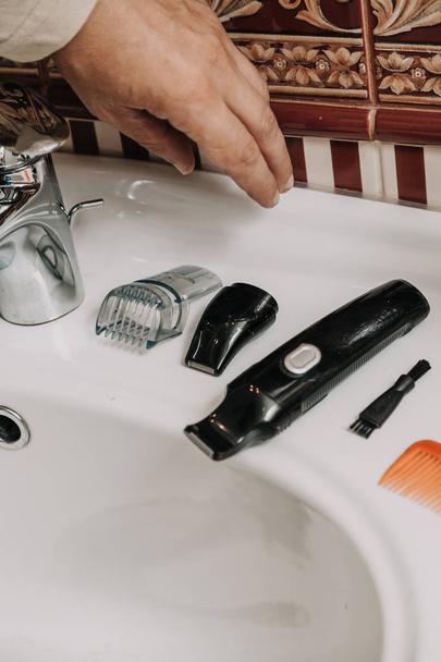 En el lavabo de azulejos es un trimmer y un conjunto de cortadores de pelo. La mano busca el recorte
 - Foto, Imagen